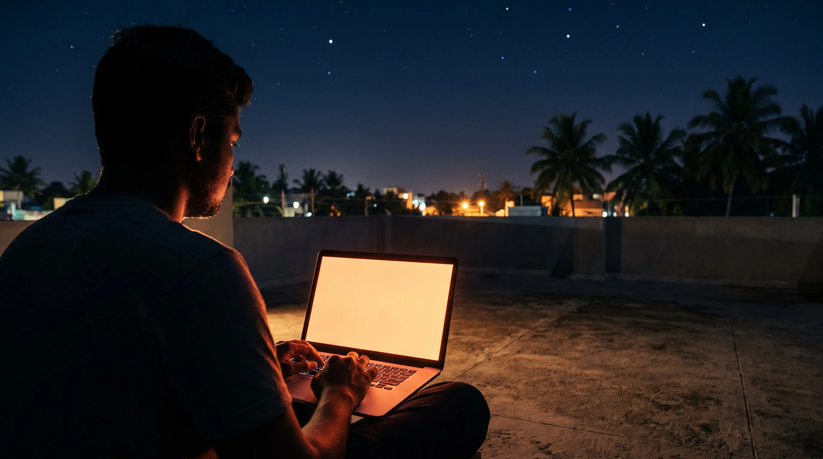 Night on a rooftop in Rajapalayam: a developer at a laptop whose screen glows orange